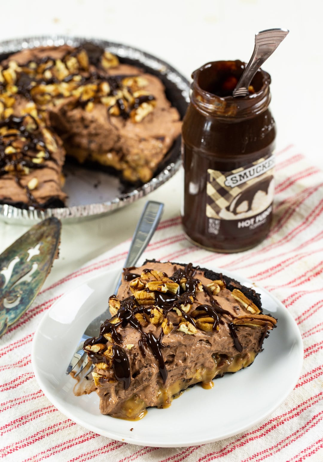Slice of pie on a plate with rest of pie and jar of fudge sauce in background.