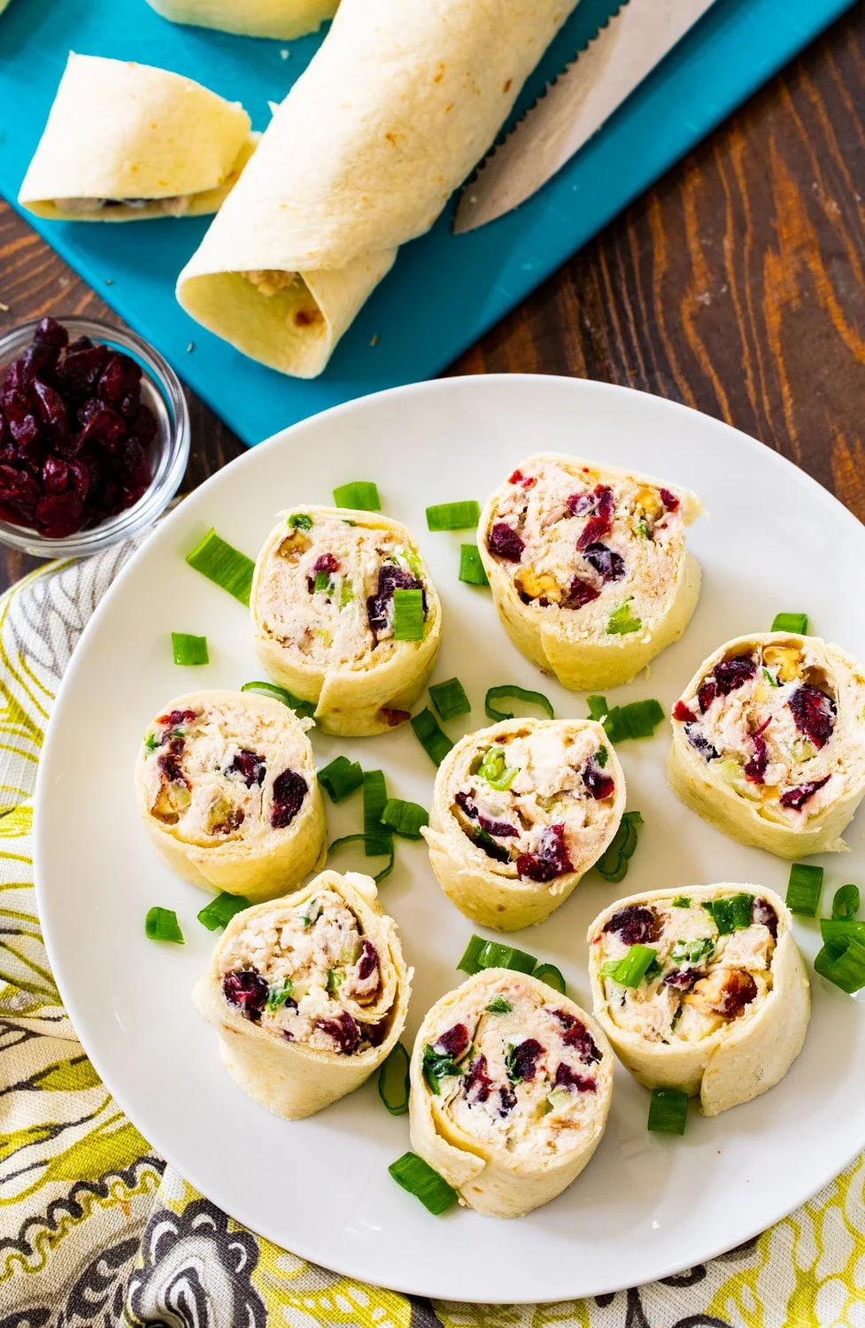 Cut pinwheels on a plate with uncut tortilla rolls on a cutting board.