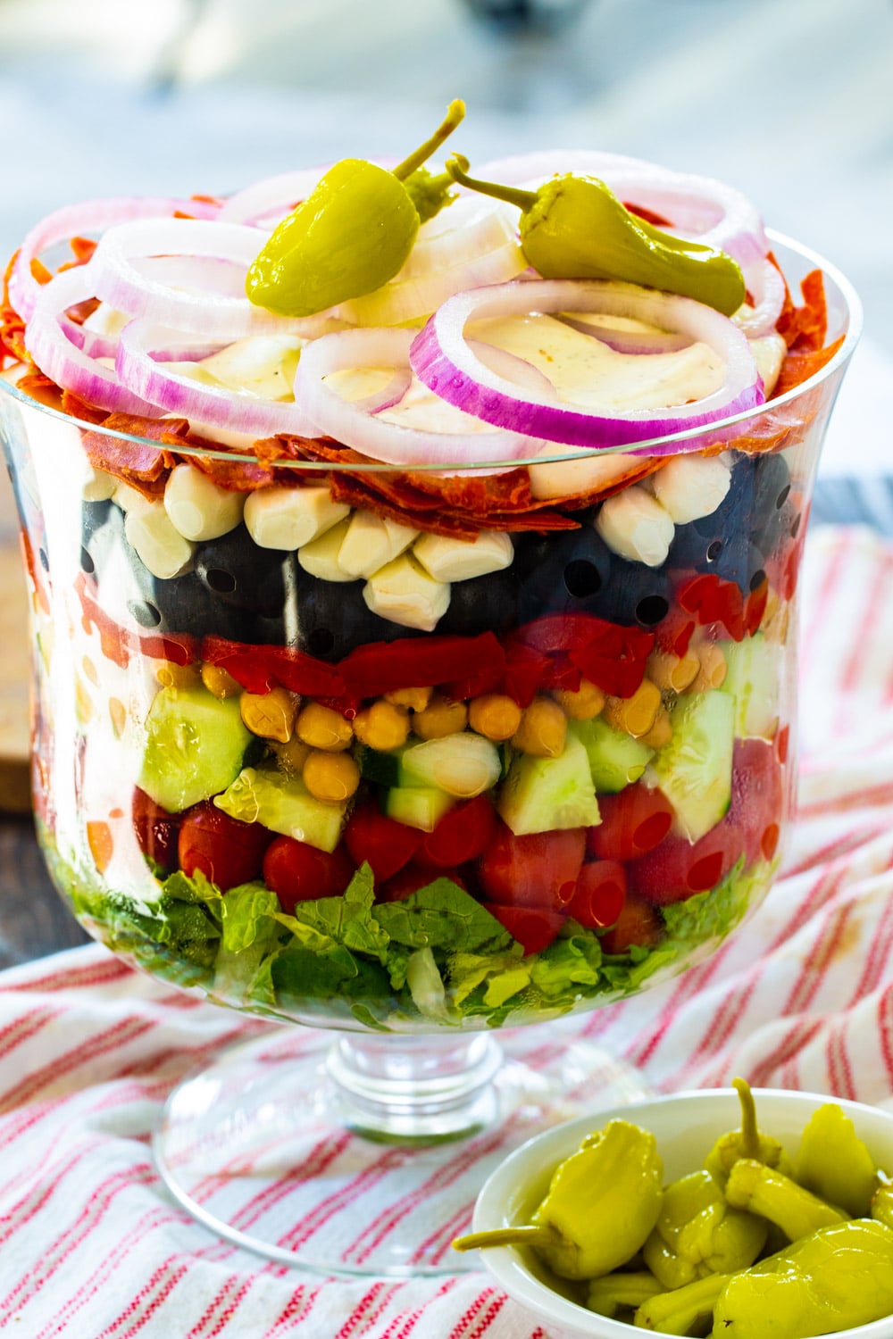 Italian Layered Salad in a glass trifle bowl with a small bowl of pepperoncini peppers in front.