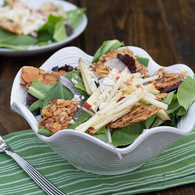 Green Salad topped with apple and almond brittle in a white bowl.