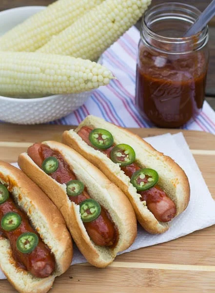 Three Hot Dogs with Dr Pepper Barbecue Sauce with corn in background.