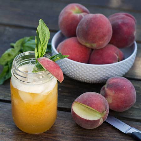 Peach Julep with basil in a mason jar surrounded by fresh peaches.