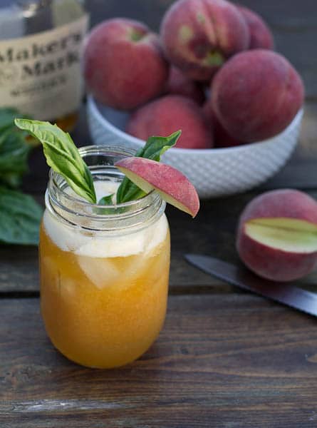 Peach Basil Julep in mason jar with fresh peaches and a bottle of bourbon.