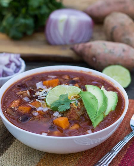 Chipotle Pork Posole in a white bowl with red onion and sweet potatoes in background.